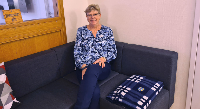 Eva Olsson sitter på en blå soffa. Hon har benen i kors och ler mot kameran. Bredvid sig har hon en blåvit filt.