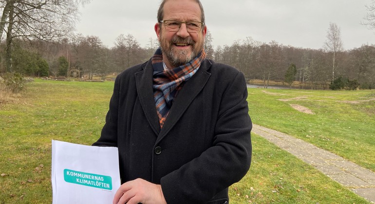 Gunnar Andersson håller i ett häfte där det står klimatmål 2030 på. Bilden är tagen utomhus med äng och skog i bakgrunden. 